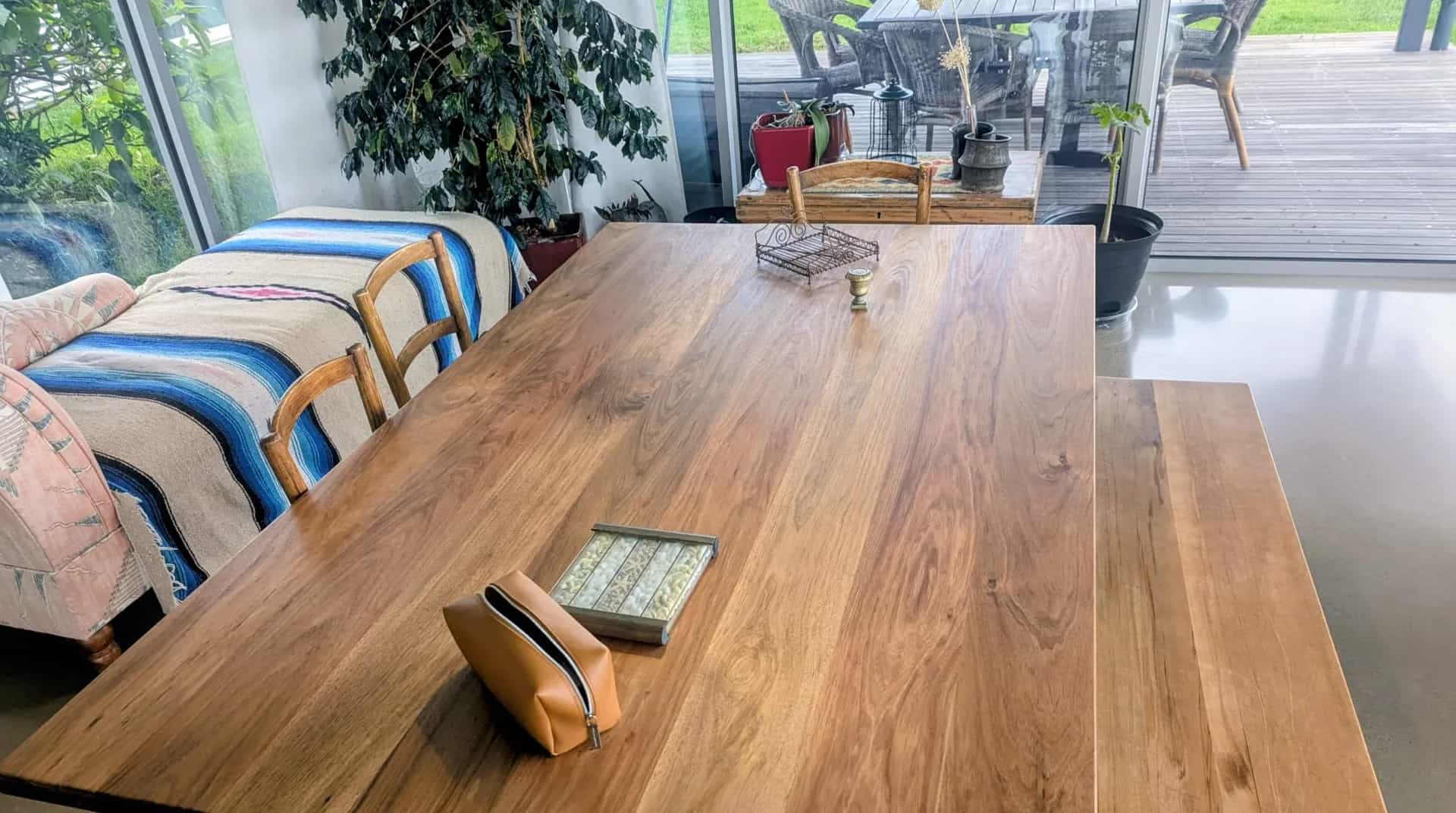 A large solid timber dining table with a matching bench in a light-filled room, looking out to a garden